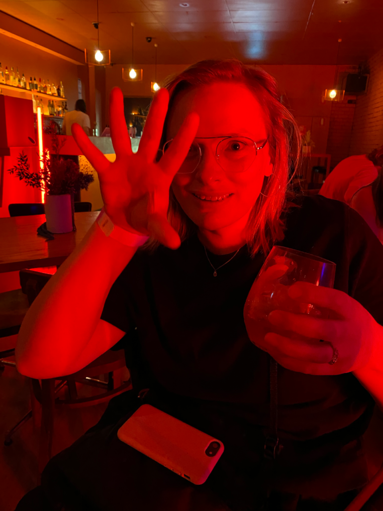 Jo at Colour, a music venue, under red light with their hand in front of their face.